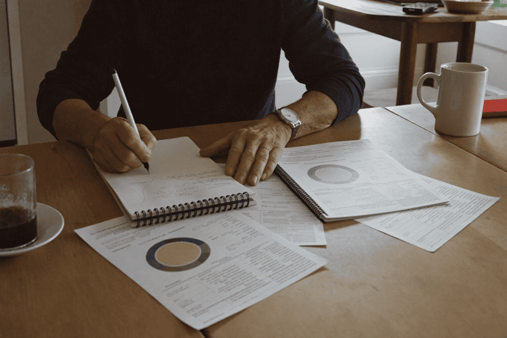 Person at kitchen table reviewing bank statements with a notepad, calculating numbers by hand — grounded, not glamorous