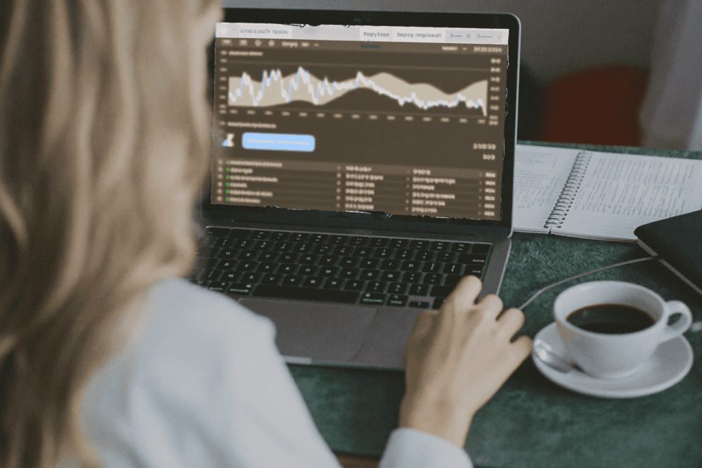 A documentary-style, over-the-shoulder photograph of a modern workspace. A laptop screen displays a complex stock trading dashboard filled with candlestick charts, real-time financial data, and account signup flow elements. Soft natural window light illuminates the scene, emphasizing realistic textures. A casual cup of coffee and a notebook are slightly out of focus in the foreground.